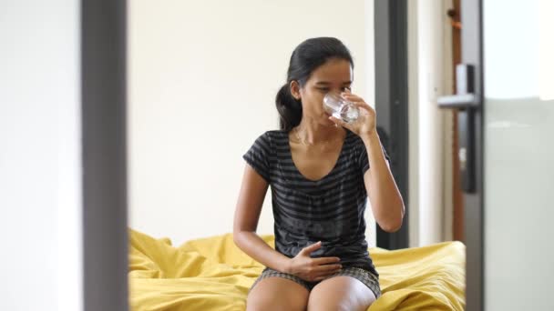 Молодая беременная азиатка сидит на кровати, прикасается к больному желудку, пьет таблетки — стоковое видео