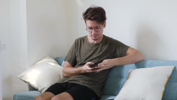 Ung moderne fyr kommunikerer i telefonen, mens du sidder i rummet på sofaen. Glad mand ringe til telefonen indendørs – Stock-video