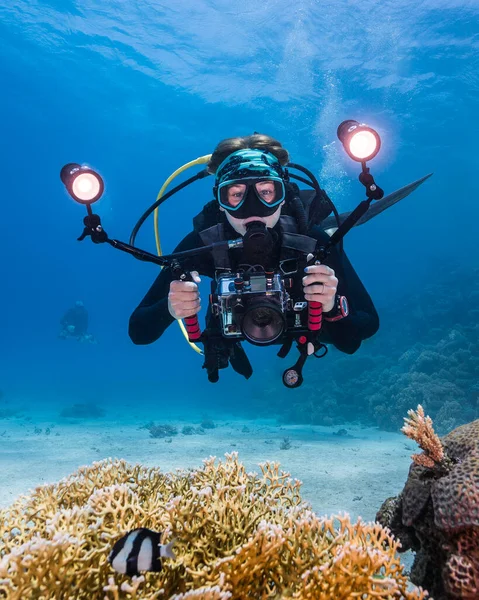 Close up of a scuba diver holding an underwater camera with two lights facing the camera with coral reef in the foreground