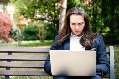 Genç bir adam bankta oturmuş dizüstü bilgisayarına danışıyor.