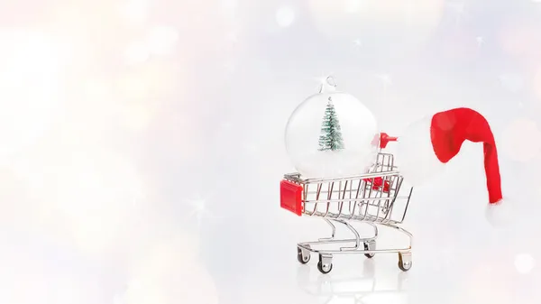 Chariot de supermarché avec boule de verre de nouvelle année et chapeau de Père Noël sur un fond de lumière.Soft focus.Christmas, fond de Nouvel An. Boule de verre de Noël avec un arbre de Noël miniature. — Photo