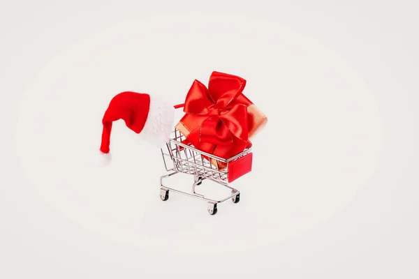 Cadeau du Nouvel An dans une boîte avec un arc rouge dans un chariot de supermarché avec un chapeau de Père Noël sur un fond clair. — Photo