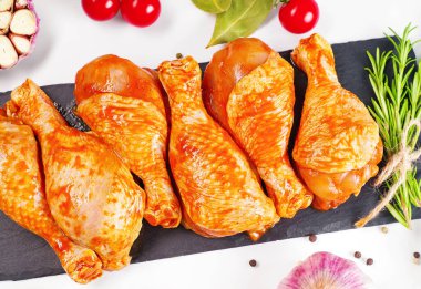 Barbekü soslu tavuk dramsticks siyah mutfak tahtası, taze biberiye yapraklı beyaz arka plan ve kiraz domatesleri, çeşitli baharatlar, yakın plan. Yarı bitmiş ürün, çabuk pişirme..