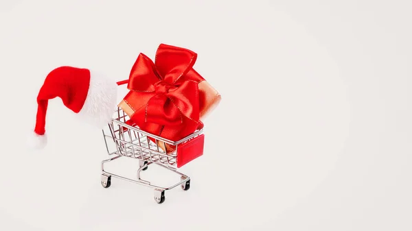 Presente de Ano Novo em uma caixa com um arco vermelho em um carrinho de supermercado com um chapéu de Papai Noel em um fundo claro.Natal e Ano Novo background.Greeting cartão, banner, poster.copy space.Glitter fundo. — Fotografia de Stock