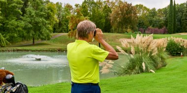 Golfçü, lazer mesafe buluculu bir golf sahasında bayrağa olan mesafeyi doğru bir şekilde ölçer. Arka planda orman var..