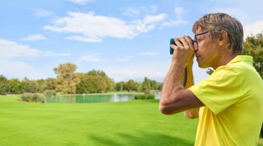 Golfçü, lazer mesafe buluculu bir golf sahasında bayrağa olan mesafeyi doğru bir şekilde ölçer. Arka planda orman var..