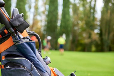 Golf arabası golf sahasında golf sopaları ve çantasıyla, yeşilin kenarına park edilmiş. Arka planda oyuncular topu cebe indirmeye hazırlanıyorlar.