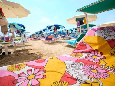 Güneşli bir yaz gününde ön planda renkli havluyla plaj yatağı. Arka planda, İtalya 'da Adriyatik Denizi kıyılarında güverte sandalyeleri ve şemsiyelerle donatılmış plaj..