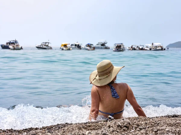 Tanned girl with a straw hat, relaxes on the beach looking at the sea on a sunny summer day, in the background the boats moored in the Gulf of Policastro, Italy.