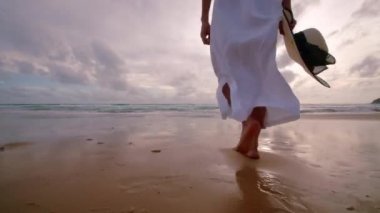 Altın gün batımında, plajda çıplak ayakla yürüyen 4K Slow motion kadın. Kumda ayak izleri bırakıyor. Yaz tatilindeki kadın turist. Tayland, Phuket plajında dinleniyor.. 