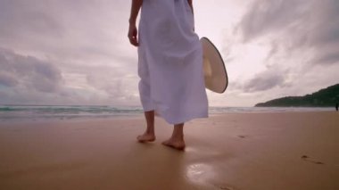 Altın gün batımında, plajda çıplak ayakla yürüyen 4K Slow motion kadın. Kumda ayak izleri bırakıyor. Yaz tatilindeki kadın turist. Tayland, Phuket plajında dinleniyor.. 