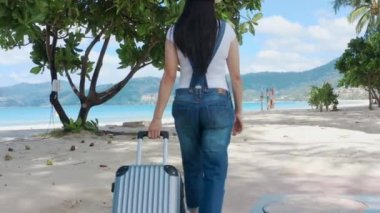 4K Asyalı gezgin kadın güneşli bir günde kumsalda yan yolda seyahat çantasıyla yürüyor. Arkadan vurulmuş. Mutlu kadın turistler plaj yolunda bavullarıyla yürürler. Seyahat tatili yazı