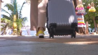 4K Asyalı gezgin kadın güneşli bir günde kumsalda yan yolda seyahat çantasıyla yürüyor. Arkadan vurulmuş. Mutlu kadın turistler plaj yolunda bavullarıyla yürürler. Seyahat tatili yazı