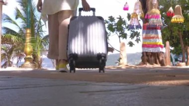 4K Asyalı gezgin kadın güneşli bir günde kumsalda yan yolda seyahat çantasıyla yürüyor. Arkadan vurulmuş. Mutlu kadın turistler plaj yolunda bavullarıyla yürürler. Seyahat tatili yazı
