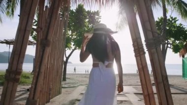 Genç turist kadınlar bambudan tropik plaja doğru yürüyorlar. Mutlu kadınlar beyaz elbise ve plaj şapkası giyer plaja yürürken rahatlarlar. Özgürlük cenneti plajı Phuket, Tayland. Yaz tatili