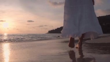 Altın gün batımında sahilde çıplak ayakla yürüyen bir kadın. Kumda ayak izleri bırakıyor. Yaz tatilindeki kadın turist. Tayland, Phuket plajında dinleniyor.. 