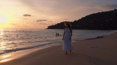 Güzel kadınların yavaş çekimde yürüdükleri beyaz elbiseler giyerler ve gün batımının tadını çıkarırlar. Günbatımı gökyüzü ile tropik plajda yürüyen turist kadınlar. Özgürlük cenneti tatil tatili yaz konsepti