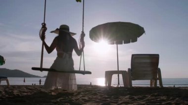 Özgürlüğün tadını çıkaran siluet görünümlü genç kadınlar gün batımına yakın salıncaklarda sallandıklarını hissediyorlar. Turist kadınlar tropik cennet sahillerinde salıncaktan hoşlanırlar. Phuket, Tayland.