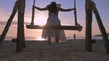 Özgürlüğün tadını çıkaran siluet görünümlü genç kadınlar gün batımına yakın salıncaklarda sallandıklarını hissediyorlar. Turist kadınlar tropik cennet sahillerinde salıncaktan hoşlanırlar. Phuket, Tayland.