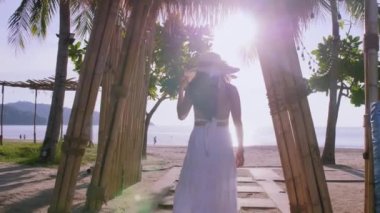 Genç turist kadınlar bambudan tropik plaja doğru yürüyorlar. Mutlu kadınlar beyaz elbise ve plaj şapkası giyer plaja yürürken rahatlarlar. Özgürlük cenneti plajı Phuket, Tayland. Yaz tatili