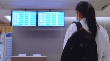 4K Asya 'lı turist kadınlar seyahat çantası takılı koruyucu fack maskesi takıyorlar. Hava alanı COVID-19 Coronavirus salgınında uçuş takvimlerini kontrol ediyorlar. Güvenli seyahat ve turizm