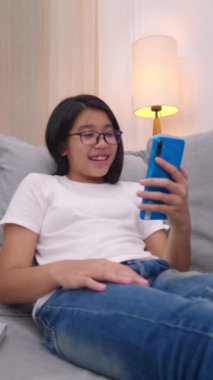 Oturma odasındaki koltukta oturan mutlu Asyalı kız cep telefonuyla ebeveyniyle konuşuyor, gülümseyen tatlı kız akıllı telefonuyla video kullanıyor ailesiyle konuşuyor. İnternet Wi-Fi üzerinden.