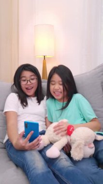 Mutlu kardeşler, oturma odasında kanepede oturan çocuklar, aileleriyle cep telefonunda konuşan, gülümseyen büyük kız, akıllı telefondan küçük kız kardeş videosu izleyip aileleriyle konuşuyorlar. 