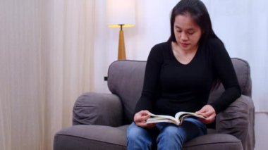 Oturma odasında kitap okuyan genç bir kadın. Kadınlar evdeki kanepede oturup kitap okuyorlar. Kadın hafta sonları boş zamanlarında rahat bir kanepede rahatlar.