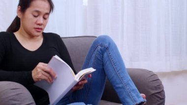 Oturma odasında kitap okuyan genç bir kadın. Kadınlar evdeki kanepede oturup kitap okuyorlar. Kadın hafta sonları boş zamanlarında rahat bir kanepede rahatlar.