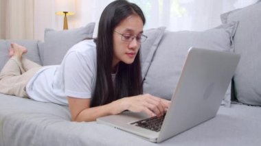 Asyalı kadın evde kanepede dinleniyor ve dizüstü bilgisayar kullanıyor. Evdeki kanepede dizüstü bilgisayar kullanan gülümseyen kadın. Kadınlar oturma odasında bilgisayarla çalışıyor.