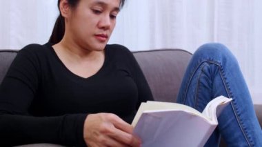 Oturma odasında kitap okuyan genç bir kadın. Evde oturan ya da kanepede uzanan ve kitap okuyan bir kadın. Kadın hafta sonları boş zamanlarında rahat bir kanepede rahatlar.