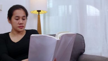 Oturma odasında kitap okuyan genç bir kadın. Evde oturan ya da kanepede uzanan ve kitap okuyan bir kadın. Kadın hafta sonları boş zamanlarında rahat bir kanepede rahatlar.