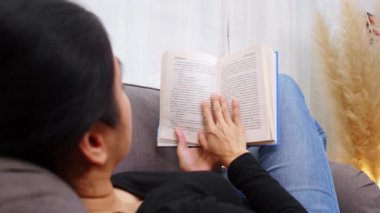 Oturma odasında kitap okuyan genç bir kadın. Evde oturan ya da kanepede uzanan ve kitap okuyan bir kadın. Kadın hafta sonları boş zamanlarında rahat bir kanepede rahatlar.