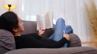 Oturma odasında kitap okuyan genç bir kadın. Evde oturan ya da kanepede uzanan ve kitap okuyan bir kadın. Kadın hafta sonları boş zamanlarında rahat bir kanepede rahatlar.