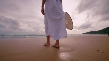 Altın gün batımında plajda çıplak ayakla yürüyen 4K kadın. Kumda ayak izleri bırakıyor. Yaz tatilinde bayan turistler Phuket, Tayland 'da dinleniyor..