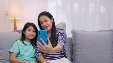 Mutlu Asyalı aile annesi ve kızı kanepede oturup akıllı telefonla evdeki oturma odasında video konferansını konuşuyorlar. Anne ve şirin küçük kız video görüşmesi. Akıllı telefondan. Teknoloji yaşam tarzı.
