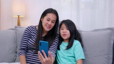 Mutlu Asyalı aile annesi ve kızı kanepede oturup akıllı telefonla evdeki oturma odasında video konferansını konuşuyorlar. Anne ve şirin küçük kız video görüşmesi. Akıllı telefondan. Teknoloji yaşam tarzı.