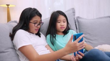 Mutlu kardeşler, oturma odasında kanepede oturan çocuklar, aileleriyle cep telefonunda konuşan, gülümseyen büyük kız, akıllı telefondan küçük kız kardeş videosu izleyip aileleriyle konuşuyorlar.