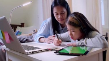 4K Asyalı küçük kız annesiyle çalışmak için dizüstü bilgisayar kullanıyor. Öğrenci çocuk ödev kitabı yazıyor. Kız evde çalışmak için bilgisayar kullanıyor. Eğitim ev okulu kavramı.