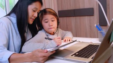 4K Asyalı küçük kız annesiyle çalışmak için dizüstü bilgisayar kullanıyor. Öğrenci çocuk ödev kitabı yazıyor. Kız evde çalışmak için bilgisayar kullanıyor. Eğitim ev okulu kavramı.