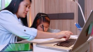4K Asyalı küçük kız annesiyle çalışmak için dizüstü bilgisayar kullanıyor. Öğrenci çocuk ödev kitabı yazıyor. Kız evde çalışmak için bilgisayar kullanıyor. Eğitim ev okulu kavramı.