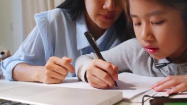 4K Asyalı küçük kız annesiyle çalışmak için dizüstü bilgisayar kullanıyor. Öğrenci çocuk ödev kitabı yazıyor. Kız evde çalışmak için bilgisayar kullanıyor. Eğitim ev okulu kavramı.