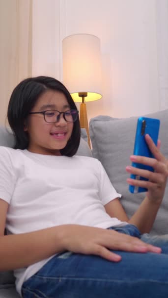 リビングルームのソファに座っている幸せなアジアの女の子は 携帯電話で親と話し 笑顔かわいい女の子は家族と話すスマートフォンでビデオ通話を使用しています インターネットWifiを通じて — ストック動画