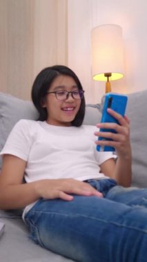Oturma odasındaki koltukta oturan mutlu Asyalı kız cep telefonuyla ebeveyniyle konuşuyor, gülümseyen tatlı kız akıllı telefonuyla video kullanıyor ailesiyle konuşuyor. İnternet Wi-Fi üzerinden.