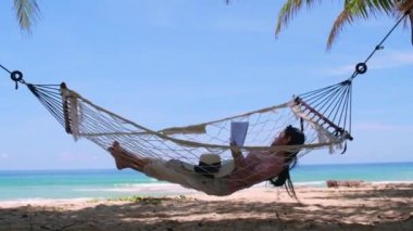 Phuket Tayland. Gün batımında sahilde hamak üzerine kitap yazan Asyalı genç bir kadın. Genç ve çekici bir kadın hamakta sallanıyor. Yaz tatili