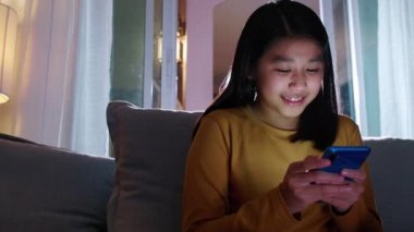 Oturma odasındaki koltukta oturan mutlu Asyalı kız cep telefonuyla ebeveyniyle konuşuyor ya da sohbet ediyor, gülümseyen tatlı kız akıllı telefondan ailenle konuşuyor. İnternet Wi-Fi üzerinden.