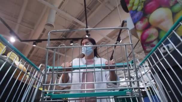 保護フェイスマスクを身に着けているアジアの女性は スーパーマーケットでいくつかの食品を選択します女性の低角度ビューCovid 19コロナウイルス発生後のスーパーマーケットで新しい通常のショッピングカートの購入材料 — ストック動画