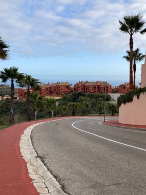 Uzakta bir deniz manzarası, palmiye ağaçları ve ön planda kırmızı bir trotuar karşısında kırmızı İspanyol kentleşmesi.