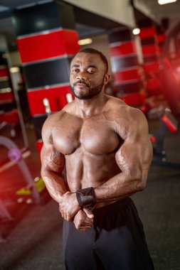 Afro-Amerikan erkek fitness eğitmeninin portresi. Vücut geliştirici pazılarını gösteriyor ve spor salonunun arka planına bakıyor.