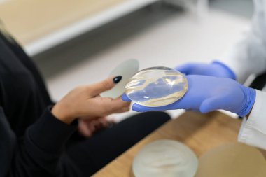 Doktor göğüs implantlarını hastanın yanında tutuyor. Kadın göğüs estetik cerrahisi kliniğini ziyaret et. Tecrübeli bir cerraha danışıyorum. Kadın göğüs implantı seçimi